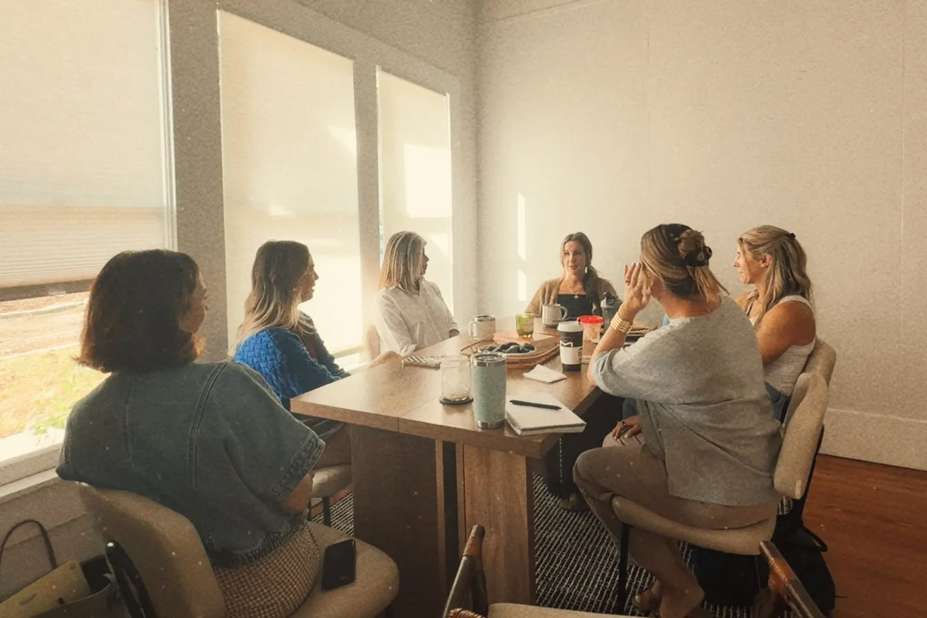 Six women gathered around a wooden table in a sunlit room, deep in conversation