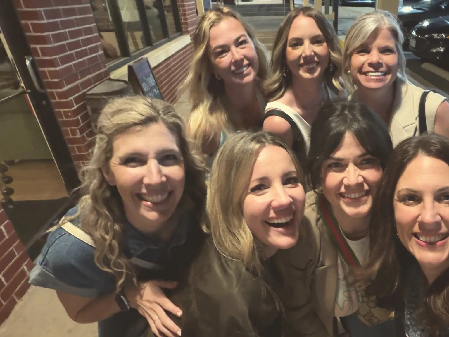 Seven friends crowded together laughing in a sidewalk selfie at night