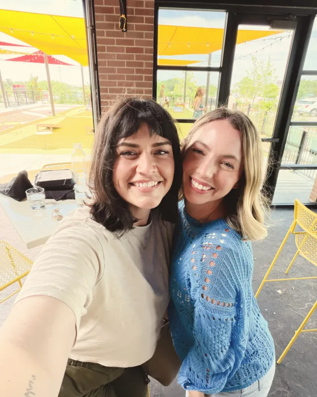 Two friends pressed cheek to cheek, grinning in a bright café